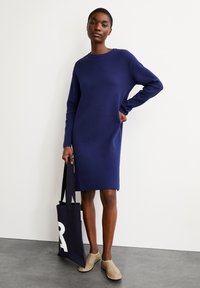 Robe pull bleu marine à manches longues et encolure ronde, arborant une coupe décontractée. La modèle porte un sac fourre-tout noir avec la lettre blanche "R."
