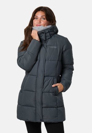 Une femme présente un manteau matelassé gris foncé à col montant doublé de polaire, qu’elle tient près de son menton sur un fond blanc uni.