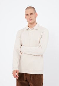 Polo a maniche lunghe beige con colletto e chiusura a tre bottoni. Realizzato in materiale morbido, presenta polsini e orlo a coste.