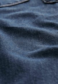 Denimová tkanina v hluboké indigo barvě s jemnou strukturou. Obsahuje viditelný vzor tkaní a mírný lesk, který ukazuje přirozené opotřebení a záhyby.