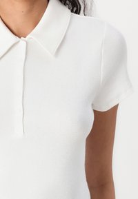 Vit kortärmad polo skjorta i mjukt stickat material, med en klassisk krage och tre knappar i placket med platt detaljering.