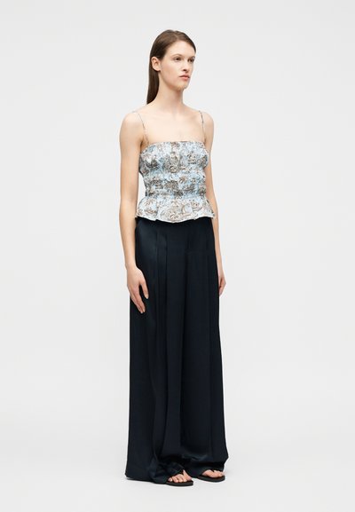 Γυναίκα που φοράει μπλε τοπ με μοτίβο και τιράντες σπαγγέτι, μαύρο παντελόνι με φαρδιά πόδια και μαύρα σανδάλια, στέκεται μπροστά σε λευκό φόντο.