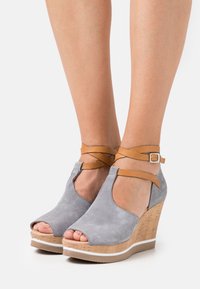 Sandales compensées en suède gris avec orteils ouverts, brides de cheville marron et bases en liège. Présentent une texture douce et une semelle blanche contrastante.