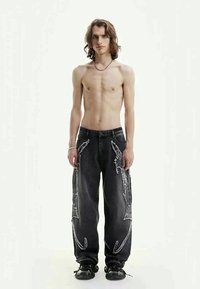 Jovem com cabelo comprido, sem camisa, vestindo calças jeans largas pretas com desenhos brancos e tênis pretos, em pé contra um fundo simples.