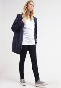 Parka azul-marinho com capuz forrado de pelúcia sintética, fecho com zipper e bolsos, combinado com uma blusa branca e jeans justos escuros, juntamente com ténis cinzentos.