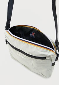 Μικρή ανοιχτόγκρι τσάντα ώμου με μαύρο ρυθμιζόμενο ιμάντα, μπροστινή τσέπη με φερμουάρ, η κύρια θήκη ανοιχτή που αποκαλύπτει μαύρο εσωτερικό με το λογότυπο της μάρκας.