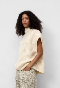 Beżowy, dziany, bezrękawnik o wysokim kołnierzu, z wzorem falistym. Połączony z wzorzystymi, jasnoszarymi spodniami.