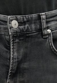 Κοντινό πλάνο της ζώνης της μαύρης τζιν παντελονιού που δείχνει μεταλλικό κουμπί με ανάγλυφο κείμενο του εμπορικού σήματος και λεπτομέρεια θηλιάς ζώνης.