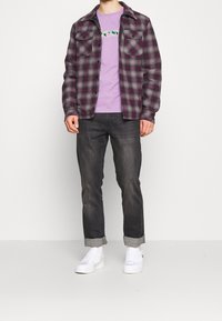 Flanelová košile s fialovo-šedým kostkovaným vzorem, světle fialové tričko, tmavé džíny a bílé tenisky. Design se dvěma kapsami, volný střih, ohrnuté manžety.