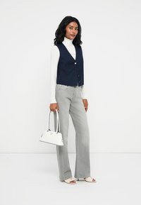 Granatowa kamizelka z pięcioma guzikami na białym golfie, szare dzwony, oraz mała biała torebka z czarnymi akcentami. Płaskie sandały.