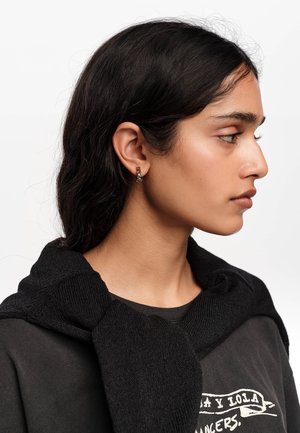 Młoda kobieta z ciemnymi włosami, ukazana z profilu, nosząca małe, kolorowe kolczyki koła, czarny sweter zarzucony na ramiona oraz ciemny t-shirt z nadrukiem.