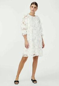 Vestido branco de renda com recortes florais, ajuste solto e mangas de três quartos, apresentando um decote redondo e uma bainha com bordas recortadas. Sandálias pretas de slide.