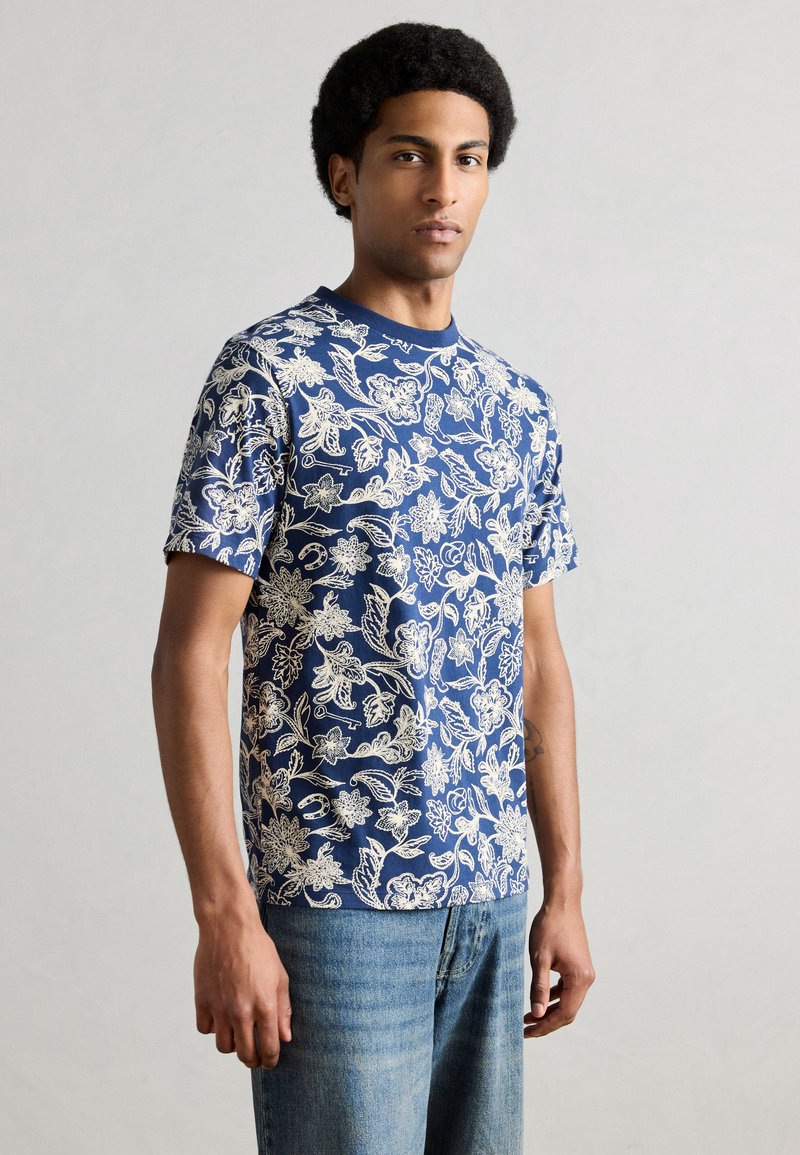 Μπλε κοντομάνικο t-shirt με λουλουδάτο σχέδιο, που παρουσιάζει περίπλοκα λευκά λουλουδάτα μοτίβα, λαιμόκοψη τύπου crew, σε συνδυασμό με ανοιχτόχρωμα τζιν παντελόνια.