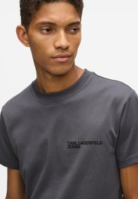 Młody mężczyzna ubrany w ciemnoszary T-shirt Karl Lagerfeld Jeans, patrzy w lewo na tle jednolitego białego tła.
