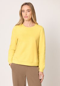 Femme debout portant un sweat-shirt jaune à manches longues et un pantalon marron clair, les mains partiellement dans les poches, sur un fond uni clair.