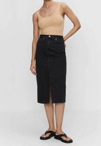Černá džínová midi sukně s předním rozparkem, vysokým pasem a klasickým designem s pěti kapsami, kombinovaná s světle béžovým žebrovaným tílkem a černými sandály.