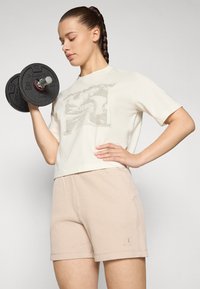 Kurzes cremfarbenes Oversized-T-Shirt mit grafischem Design und beigen, gerippten Sportshorts. Modell hält ein schwarzes Hantel.