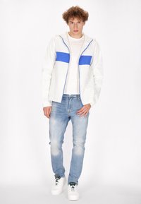 Bela zip-jacket s sivo-modro vodoravno črto, v kombinaciji z svetlo modrimi džinsom in belimi supergami. Enostaven dizajn, sproščen kroj.