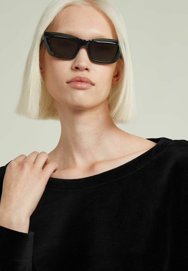 Schwarze Cat-Eye-Sonnenbrillen mit dicken Rahmen, dunklen Gläsern und glänzender Oberfläche, getragen von einer Person mit kurzen, glatten, platinblonden Haaren.