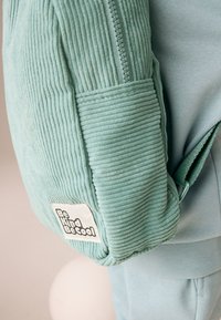 Veste en velours côtelé bleu clair à surface texturée, avec une fermeture éclair et une étiquette blanche "Be Kind Be Cool" sur la poche.