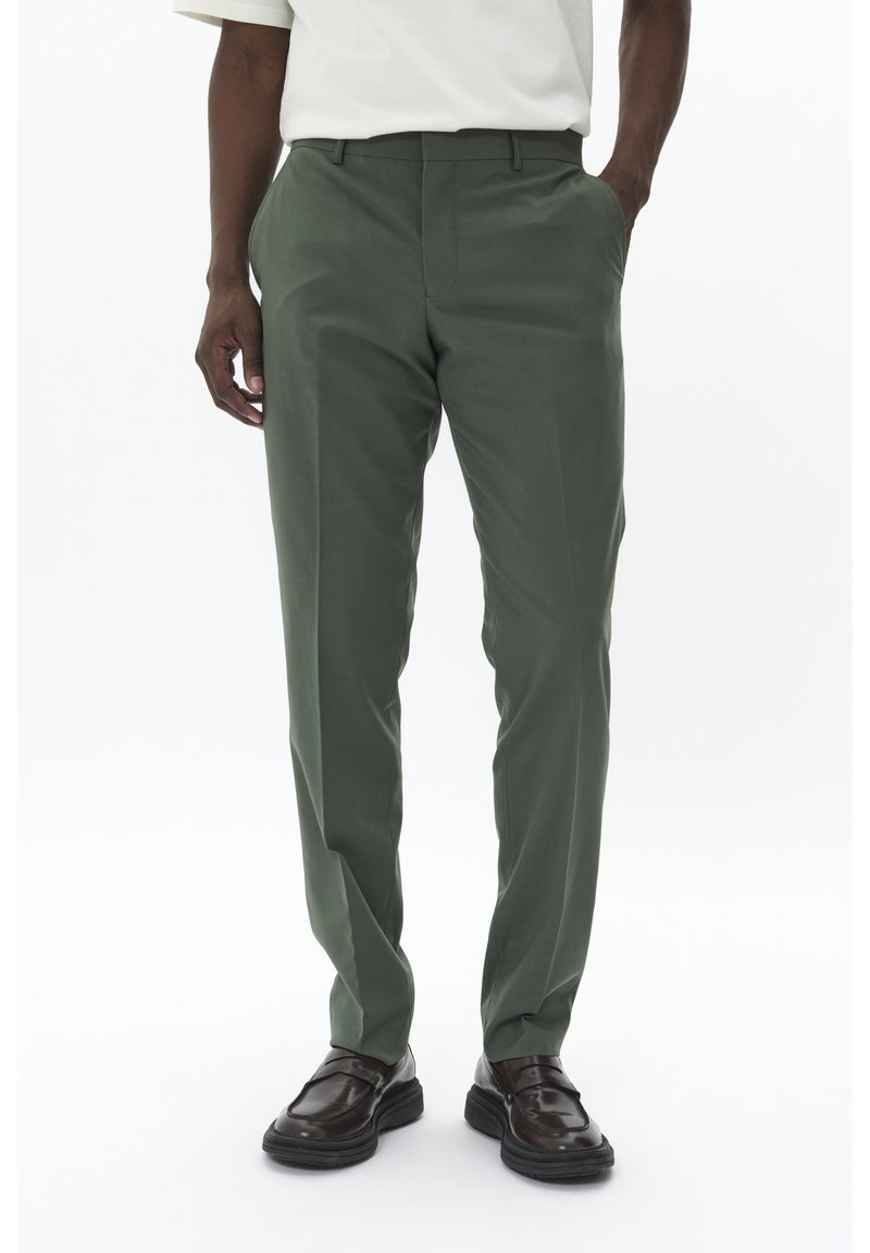 Pantalons vert olive à coupe ajustée, dotés de poches latérales et d'une texture lisse, associés à des chaussures marron foncé.