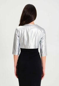 Zilveren cropped jacket met driekwart mouwen, met een gladde, glanzende textuur. Gepaard met een aansluitende zwarte jurk eronder.