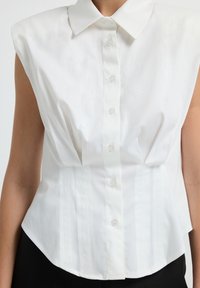 Mouwloze witte blouse met knopen en een puntige kraag, gedragen over zwarte broek, afgebeeld van halverwege de torso tot de nek.