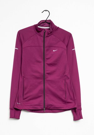 Chaqueta deportiva morada de Nike con cremallera, cuello alzado, bolsillos con cremallera y franjas reflectantes en las mangas, colgada en una percha negra.