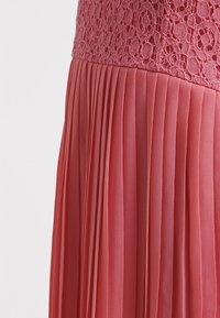 Plisovaná sukně v jemné korálové barvě, která má texturovanou krajkovou korzetovou část a splývavou, lehkou látku.