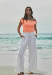Top arancione luminoso senza spalle abbinato a pantaloni bianchi in lino larghi con tasche. In piedi su una spiaggia con acqua sullo sfondo.