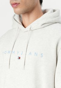 Lys grå hettegenser laget av myk tekstil, med blå "TOMMYJEANS" broderi og et lite logo med røde og blå detaljer. Justerbare snorer.