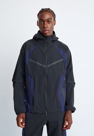 Black and navy athletic jacket with a hood, featuring zippered pockets, a sleek design, and reflective details on the chest and sleeves.