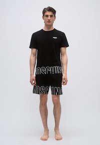 Νέος άνδρας που στέκεται ξυπόλητος, φοράει μαύρο t-shirt Moschino και ταιριαστό σορτς με μεγάλα λευκά γράμματα λογότυπου στο κάτω μέρος.