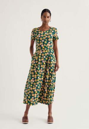 Kvinde står iført en grøn midi-kjole med korte ærmer og blomstermønster i gult og pink samt brune slip-on træsko.