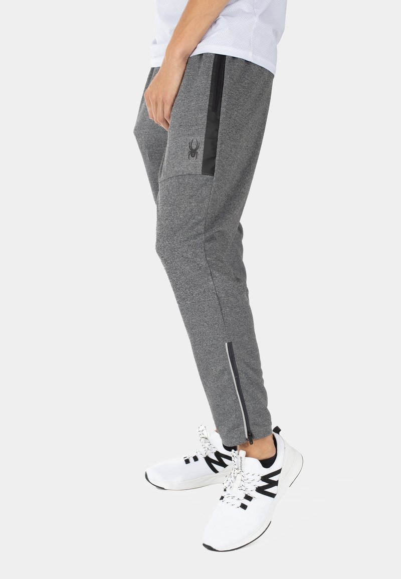 Homme portant un pantalon de jogging gris avec une fermeture éclair à la cheville et un logo araignée noir, accompagné de chaussures de sport blanches avec des accents noirs.