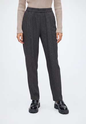 Personne portant un pantalon gris foncé sur mesure, un haut côtelé à manches longues beige et des mocassins noirs brillants et épais, debout sur un fond blanc.