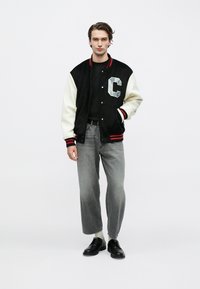 Νεαρός άνδρας που στέκεται φορώντας μαύρο varsity jacket με λευκά μανίκια, γκρι τζιν, άσπρες κάλτσες και μαύρα επίσημα παπούτσια σε λευκό φόντο.