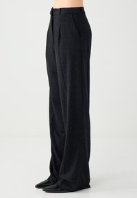 Personne portant un pantalon gris foncé taille haute à jambes larges avec poches latérales et chaussures noires à bouts pointus, sur un fond clair uni.