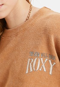 Skarżonoczerwony, teksturowany sweatshirt z okrągłym dekoltem. Wyróżnia się niebieskim i białym haftem z napisem "SŁOŃCE WSTANIE ROXY."