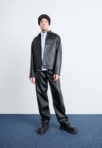 Veste en simili cuir noir avec une fermeture éclair, poche latérale, portée sur une chemise blanche. Un pantalon en simili cuir noir et des chaussures noires épaisses complètent le look.