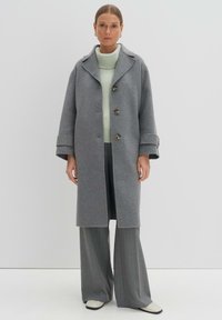 Cappotto di lana grigio con colletto a pistagna e cinque bottoni, indossato sopra un maglione a coste di un verde chiaro e pantaloni grigi a righe.