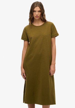Vrouw met golvend haar in een olijfgroene, kort mouwen, midi-lengte t-shirt jurk die tegen een eenvoudige witte achtergrond staat.