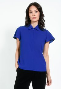 Kongeblå kortærmet bluse med lynlåsfortil, spidse kraver og afslappet pasform. Fremstillet af glat stof med en let glans.