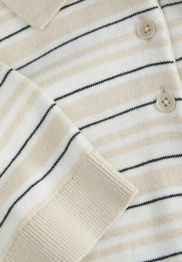 Sweter polo w kolorze kremowym z beżowymi i czarnymi paskami, z klasycznym kołnierzykiem, czterema guzikami oraz ściągaczami na rękawach dla uzyskania teksturowanego wykończenia.