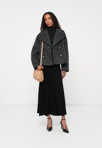 Giacca doppiopetto in misto lana grigia con grandi bottoni, abbinata a una gonna nera plissettata. Tacchi a spillo neri e una borsa beige completano il look.