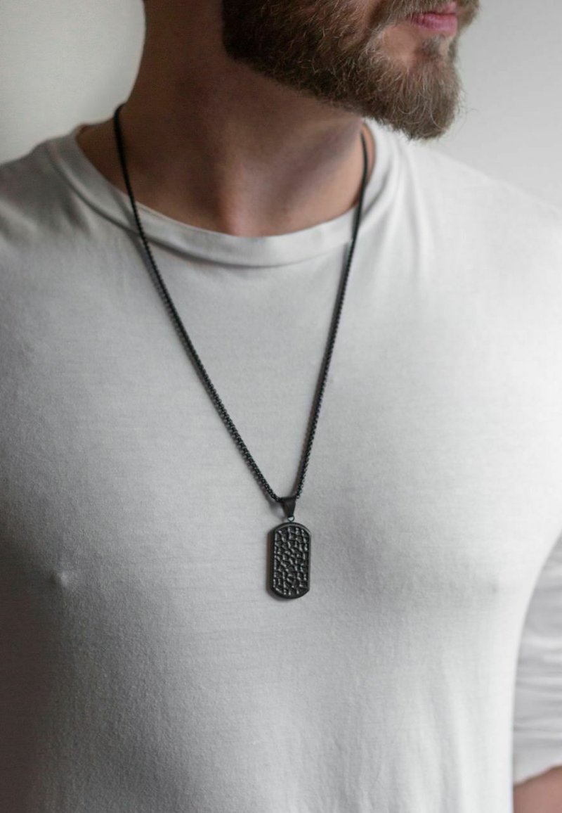 Schwarzes Hundemarken-Pendant an einer Kette, mit einer strukturierten Oberfläche, über einem hellgrauen T-Shirt getragen. Einfaches, minimalistisches Design.