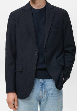 Mężczyzna ubrany w ciemnogranatową marynarkę nałożoną na granatowy, prążkowany sweter, w jasnoniebieskich dżinsach, ręce swobodnie opadające wzdłuż ciała.