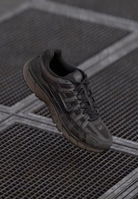 Nike tossud musta võrgusilmas tumedate hallide aktsentidega, millel on tekstureeritud talla ja silmatorkav paeltesüsteem, paigutatud ruudustiku taustale.