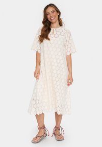 Linneklänning i off-white med blommönster, korta ärmar, rund halsringning och en knytdetalj. Bärs med färgglada, bandade sandaler.