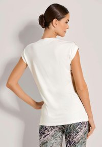 Vrouw met haar in een knot, draagt een mouwloze witte top en gestreepte broek, staat met één hand op haar heup en kijkt weg.